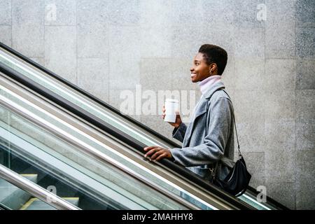 Vista laterale di un'elegante donna afro-americana in un elegante cappotto e maglione. Lei è con una tazza di caffè e una borsa sorridente e cavalcando le scale in movimento in città Foto Stock