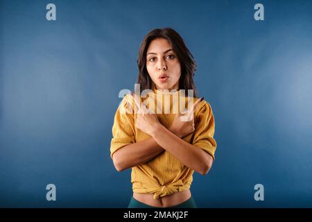 Giovane bella donna che indossa giallo top isolato su sfondo blu incrocia braccia e punti laterali, sceglie tra due opzioni. Scioccato, un incr Foto Stock