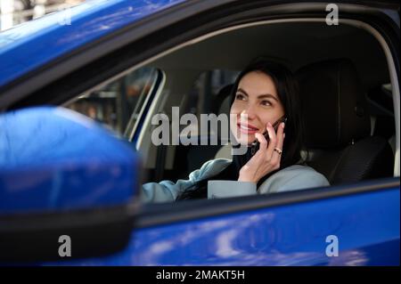 Donna attraente seduta in auto sul sedile del conducente, utilizzando smartphone, parlare con i clienti, pianificare incontri di lavoro. Foto Stock