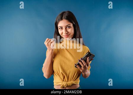 Giovane ragazza dai capelli scuri sorridente felice indossando la parte superiore gialla isolata sopra lo sfondo blu che guarda lo schermo del telefono, celebrando una vittoria in linea, overjoye Foto Stock