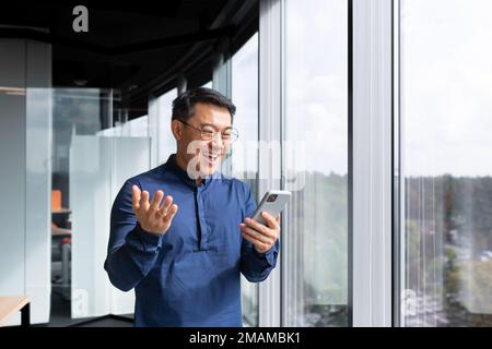 Riuscito boss asiatico vicino alla finestra ha ricevuto le buone notizie in linea, l'uomo d'affari usa il telefono e celebra la vittoria ed i buoni risultati di realizzazione all'interno dell'ufficio, l'uomo in camicia ed i vetri eccitati gioiscono. Foto Stock