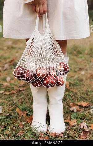 Borsa a corda con mele rosse. Vista delle gambe femminili in stivali bianchi che attraversano il giardino autunnale con borsa ecologica in mano. Concetto di zero sprechi. Nessuna plastica Foto Stock