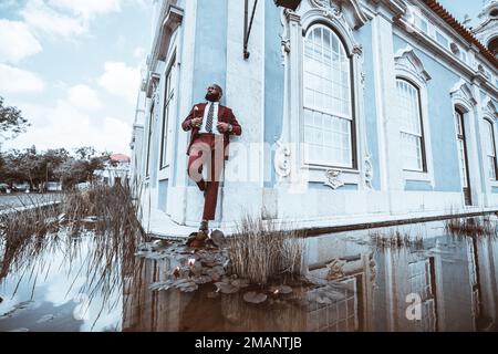 Un imponente calvo africano con una barba folta che indossa un abito sartoriale rosso, in cui sembra un drappeggio, si appoggia all'angolo di un re blu Foto Stock