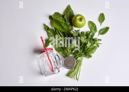 alcune verdure e una mela su una superficie bianca con una paglia rossa in alto a destra è un vaso di vetro pieno di acqua Foto Stock