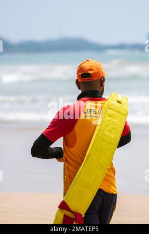 Bagnino in servizio, camminando sulla spiaggia sabbiosa mentre trasportano un tubo di salvataggio sulla spalla, bagnino vista posteriore foto. Foto Stock