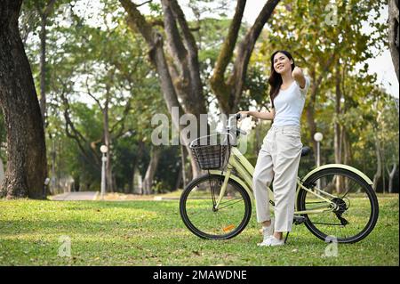 Felice e chill giovane donna asiatica in abiti casual guardando la bella vista della natura, riposandosi dopo la bicicletta nel bellissimo parco pubblico. Foto Stock