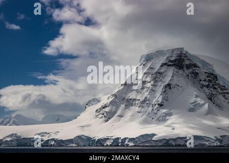 Ripida cima di montagna sulla penisola antartica. Picco coperto di fitti strati di neve e ghiaccio. Nuvole, cielo blu sullo sfondo. Foto Stock