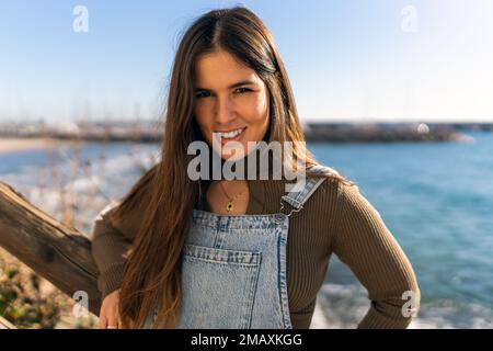 Felice femmina ispanica guardando la macchina fotografica mentre si trova in piedi sulla costa vicino al mare sereno sotto il cielo blu in estate Foto Stock