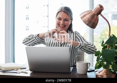 Se tonights il mio ultimo, lasci i miei capretti vedere i miei contanti. una donna d'affari matura con un gesto a forma di cuore video chattare sul suo notebook in un ufficio moderno. Foto Stock