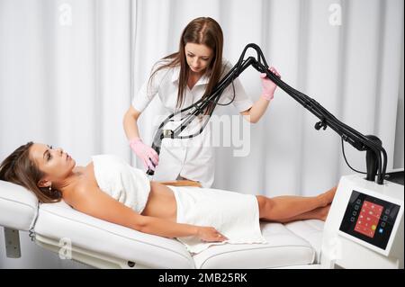 Medico cosmetologo utilizzando erbium ablative laser macchina durante l'esecuzione di procedure di sollevamento su addome femminile. Giovane donna che riceve un trattamento laser in clinica di cosmetologia o salone di bellezza. Foto Stock