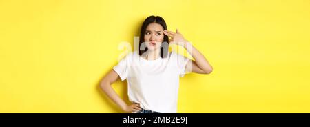 Donna che mi mostra uccidere prego gesto, sparando in testa con la pistola dito dalla noia, in piedi sconvolto su sfondo giallo Foto Stock