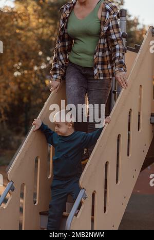 Ragazzino con sindrome di Down che scende per le scale sul parco giochi con la madre Foto Stock