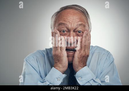 Hai appena visto quello che ho visto? un uomo d'affari anziano in piedi contro uno sfondo grigio studio con il suo volto nelle sue mani e guardando sorpreso. Foto Stock