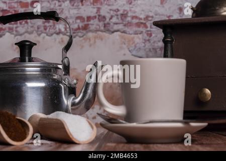 Tazza da caffè con caraffa per il latte, macinacaffè e cucchiai di legno con zucchero e caffè macinato su un tavolo di legno con parete in mattoni sullo sfondo Foto Stock