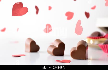 I bonbonbonbons di cioccolato a forma di cuore su un tavolo bianco e molti ritagli di cuore rosso cadono giù con fondo isolato bianco. Vista frontale. Foto Stock