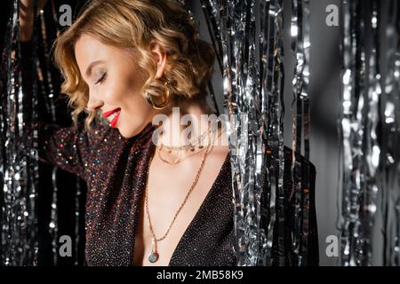 vista laterale di donna felice in vestito nero in piedi vicino lucido tenda di tinsel su grigio, immagine stock Foto Stock