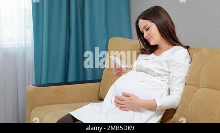 Donna incinta si siede sul divano e guarda la foto a ultrasuoni Foto Stock