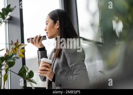 Vista laterale del designer asiatico di interni che registra messaggi vocali sullo smartphone e tiene il caffè in studio, immagine di scorta Foto Stock