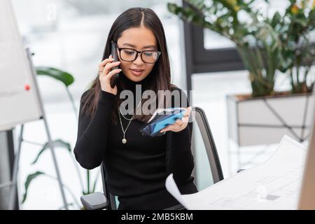 Designer asiatico sorridente che parla su smartphone e tiene campioni di colore vicino a BluePrint in ufficio, immagine stock Foto Stock