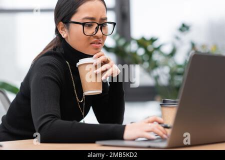 Ha sconvolto il designer asiatico tenendo la tazza di carta e utilizzando il computer portatile in ufficio, immagine di scorta Foto Stock