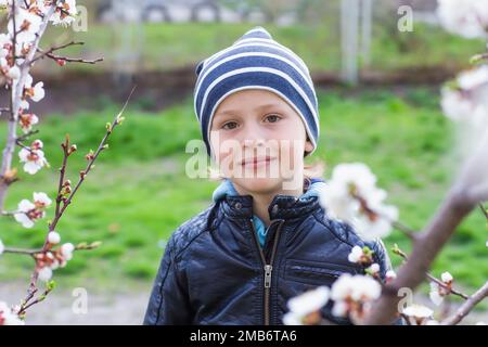 Ritratto di un ragazzino tra i fiori di ciliegio in primavera. Un bambino in una passeggiata si nasconde dietro un cespuglio. Nascondere e cercare in natura. Foto Stock