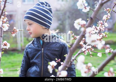 Ritratto di un ragazzino tra i fiori di ciliegio in primavera. Un bambino in una passeggiata si nasconde dietro un cespuglio. Nascondere e cercare in natura. Foto Stock