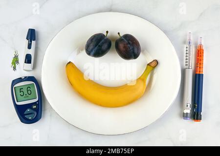 Banana e prugne sotto forma di un volto sorridente su un piatto bianco e penne per siringa per insulina e un glucometro come posate su un tavolo di marmo Foto Stock