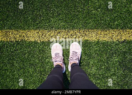 Gambe di una donna in piedi su un parco giochi di sport verde di fronte a una linea gialla, vista dall'alto Foto Stock