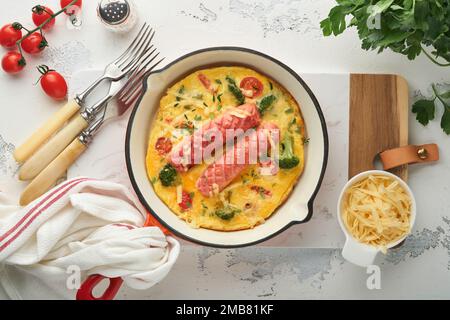 Omelette o frittata con due salsicce, formaggio fuso, pomodori ciliegini, broccoli e prezzemolo su padella rossa o teglia di ferro su fondo bianco di cemento vecchio Foto Stock