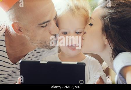 Immagine ravvicinata di due genitori che baciano la loro bambina sulle guance mentre gioca con la tavoletta (lei scopa la lingua fuori) Foto Stock