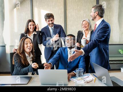 Team di colleghi maschi e femmine multirazziali felici in abiti formali che si riuniscono vicino a un computer portatile e si divertono con le notizie in un ufficio moderno Foto Stock