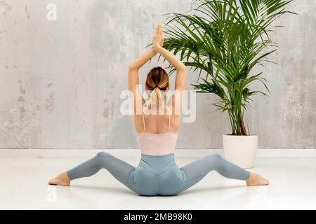 Vista integrale della schiena di una femmina a piedi nudi snella in abbigliamento sportivo seduta sul pavimento in asana yoga con braccia rialzate in studio Foto Stock