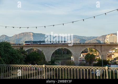 Treno elettrico bianco e arancione che attraversa un vecchio ponte ad arco a Villajoyosa, Alicante, Spagna Foto Stock