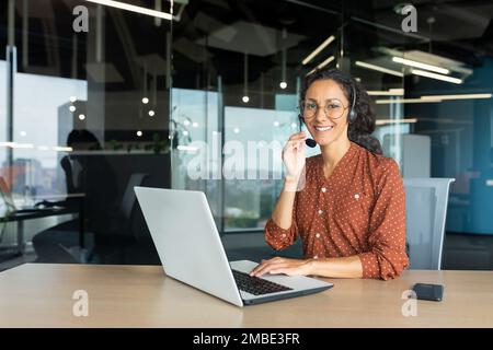 Ritratto di una giovane bella insegnante di donna latino-americana. Seduti alla scrivania in una cuffia. Insegna online, apprendimento a distanza attraverso una videochiamata da un laptop. Guarda la fotocamera con un sorriso. Foto Stock