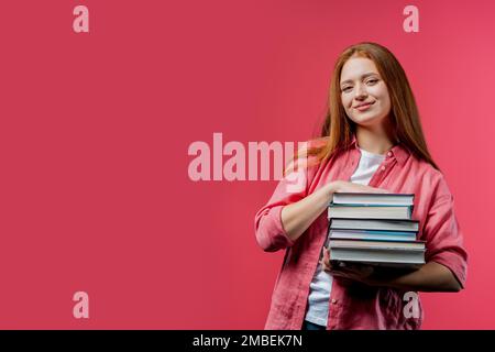 Un allievo intelligente tiene una pila di libri universitari dalla biblioteca dell'università su sfondo rosa. Felice ragazza dai capelli rossi sorride, è felice di laurearsi. Copia Foto Stock