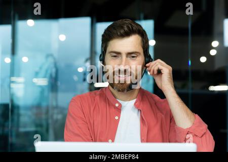 Uomo d'affari maturo di successo con cuffie per videochiamate che lavorano all'interno dell'ufficio, un uomo che si occupa di consulenza online con i clienti, che parla con i colleghi durante una riunione remota online, che si siede sul posto di lavoro con un computer portatile. Foto Stock