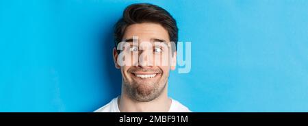 Colpo di testa di giovane uomo che fa le espressioni divertenti, sorridente e squinting, in piedi su sfondo blu Foto Stock