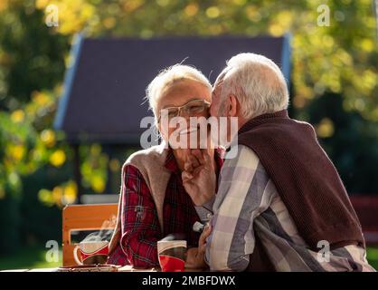 Coppia anziana seduta sulla terrazza nel cortile, bere caffè, parlare e baciare Foto Stock