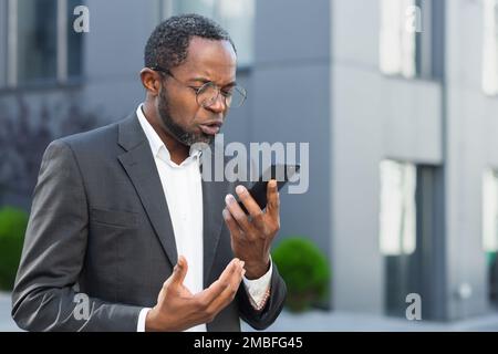 Boss arrabbiato e arrabbiato che parla e urla sul telefono cellulare, uomo d'affari afro-americano in tuta d'affari che cammina fuori ufficio e che parla con i subordinati. Foto Stock