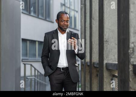 Serio afro capo americano in tuta di affari a piedi fuori ufficio edificio, uomo maturo in possesso di telefono, uomo d'affari concentrando la lettura del messaggio e la digitazione di testo. Foto Stock