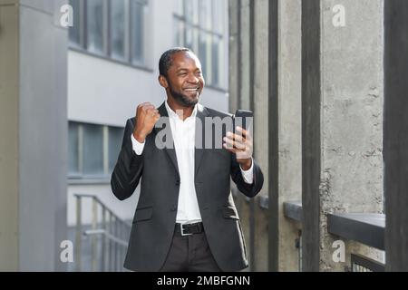 Successo african american boss fuori ufficio edificio utilizzando telefono uomo d'affari in tuta celebrando la vittoria, successo di successo, uomo che tiene la mano in su trionfo gesto, la lettura di notifica online Foto Stock