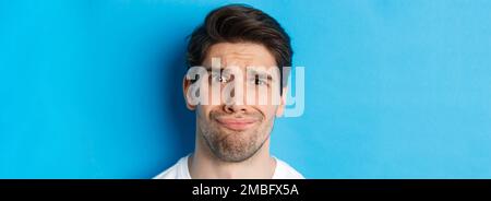 Colpo di testa di uomo scettico e deluso, grimacing complicato, in piedi su sfondo blu Foto Stock