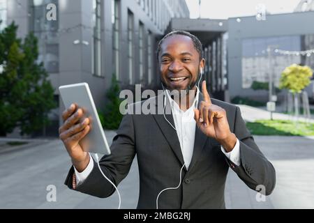 Boss afro-americano di successo fuori sede con tablet computer e cuffie parlare in remoto con i colleghi in videochiamata, uomo d'affari sorridendo e guardando la fotocamera in tuta camminare. Foto Stock