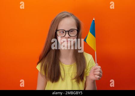 Una ragazza con sindrome di Down detiene la bandiera dell'Ucraina. sfondo arancione Foto Stock