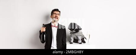 Uomo triste piangendo e bevendo champagne dalla bottiglia, mentre pug in simpatico costume da festa fissando confuso, in piedi su sfondo bianco Foto Stock