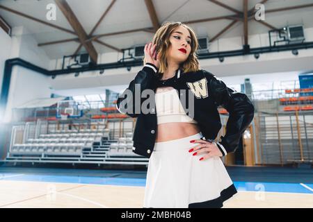 Una cheerleader che indossa una divisa bianca e nera con un ponticello nero che posa in una sala sportiva. I supporti sono sfocati sullo sfondo. Foto di alta qualità Foto Stock