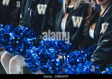 Primo piano di cheerleaders in jumper neri seduti in fila e tenendo in ginocchio pom blu e lucenti. Foto di alta qualità Foto Stock