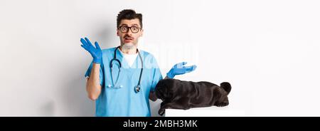 Veterinario medico intern in scrub shrugging, confuso come esaminare il cane, pug sdraiato sul tavolo, sfondo bianco Foto Stock