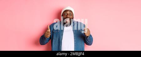 Impressionato afroamericano hipster ragazzo mostrando pollici-up, approvare e come offerta, lodando grande scelta, in piedi su sfondo rosa Foto Stock