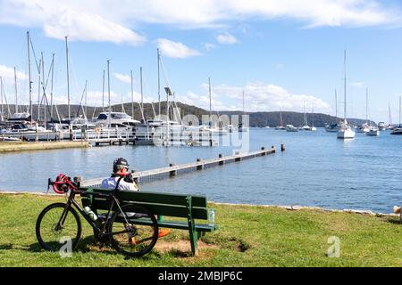 Maschio strada ciclista prende una pausa Bayview Sydney Australia sul panchina del parco godendo delle vedute attraverso Pittwater e barca marina estate 2023 Foto Stock